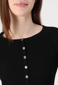Μαύρο ριμπ τοπ με στρογγυλή λαιμόκοψη και πέντε χρυσά κουμπιά μπροστά. Το ύφασμα έχει υφή και ελαστικότητα.