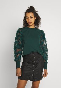 Green sweatshirt with textured sheer sleeves adorned with ruffles, paired with a black button-front mini skirt made of leather-like material.
