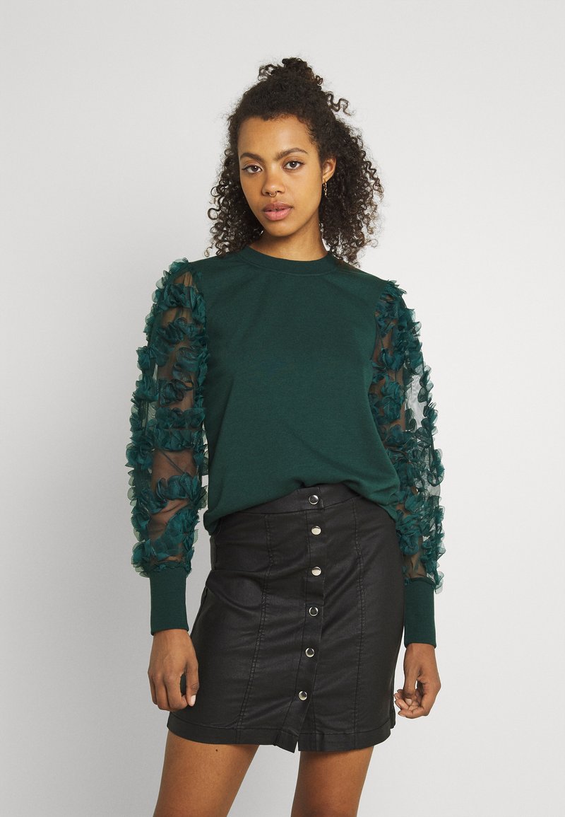 Green sweatshirt with textured sheer sleeves adorned with ruffles, paired with a black button-front mini skirt made of leather-like material.