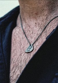 Silversmycke med ett tårformat hänge som har intrikata gravyrer, hängande från en tunn kedja, bärs mot en lätt hårig bröstkorg.