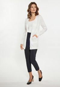 Cardigan alb cu deschidere frontală, cu dungi gri deschis, mâneci lungi și buzunare laterale, combinat cu pantaloni tapered negri și pantofi cu toc negri.