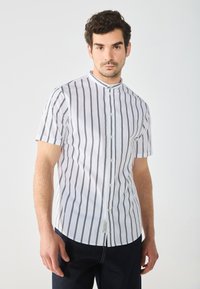 Camicia a maniche corte con colletto, di colore bianco con strisce nere verticali, dotata di chiusura a bottoni e orlo leggermente curvato.