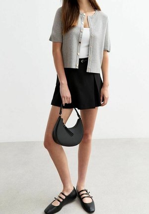 Femme portant un cardigan gris à manches courtes boutonné, un haut blanc, une jupe plissée noire, des chaussures Mary Jane noires, tenant un sac à main noir en forme de croissant.