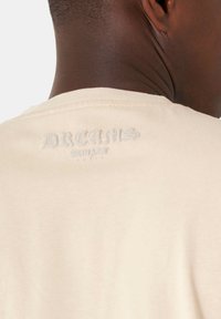 Camiseta de algodón color crema con un logo texturizado "DREAMS" bordado en el cuello trasero. Diseño de manga corta y cuello redondo.