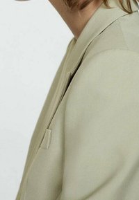 Světlý zelený blazer s přiléhavým střihem, který má klopový límec, minimalistický design a jemnou texturu. Viditelná jedna malá boční kapsa.