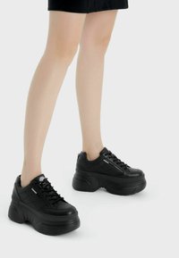 Ben iförda svarta plattformssneakers med tjocka sulor, stående på en vit bakgrund.