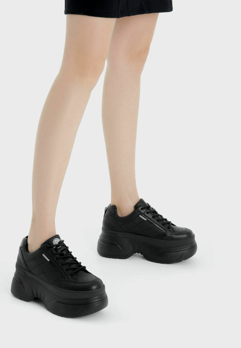 Ben iförda svarta plattformssneakers med tjocka sulor, stående på en vit bakgrund.