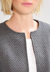 Veste grise texturée avec un col rond, présentant un motif subtil et sans fermetures, portée par-dessus un haut blanc.