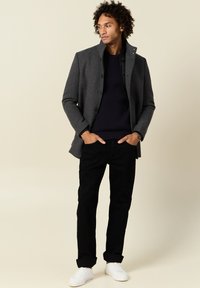 Giacca in misto lana grigia con colletto alto, maglione nero texturizzato, pantaloni neri e sneakers bianche. Il modello sta in piedi in modo informale con le mani in tasca.