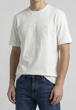 Muškarac koji nosi bijelu majicu s teksturiranim uzorkom palminog drveta i plave traperice, stoji s rukama opuštenim uz tijelo.