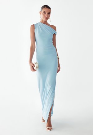 Kvinde iført en lyseblå kjole med ét skulderstrop og en slids ved låret, der holder en sølvfarvet clutch, iført hvide sandaler og guldsmykker.