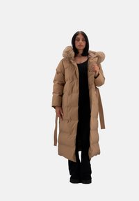 Lang brun quilted frakke med hætte i pels og detalje med snøring i taljen, samt knaplukning. Har en quiltet struktur og sidelommer.