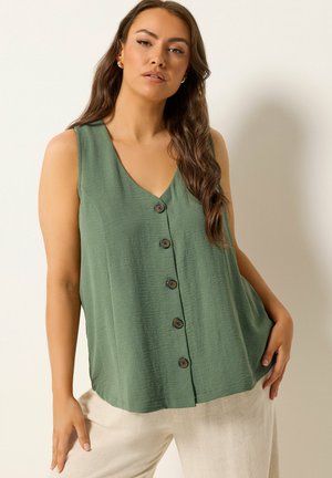 Kobieta o długich brązowych włosach, ubrana w zieloną bluzkę na guziki bez rękawów i jasnobeżowe spodnie, pozująca na tle jednolitego jasnego tła.