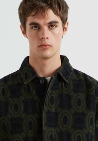 Giacca color verde e blu navy con colletto, caratterizzata da un ampio motivo ovale, texture in corduroy e chiusura a bottoni. Il tessuto presenta sottili righe.