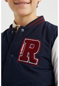 Tummansininen pommikaukkari, jossa on punainen, tekstuurilla varustettu "R"-pälkä, valkoiset hihat, ribbireunainen kaulus punaisilla ja tummansinisten raidoilla sekä napitus.