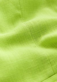 Close-up van geplooide, gestructureerde limoen-groene stof met een zichtbaar geweven patroon en natuurlijke lichtaccenten.