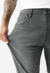 Grå denimbukser med en tætsiddende pasform, der har en talje, fem lommer og synlige syninger. En hånd trækker i stoffet ved låret.