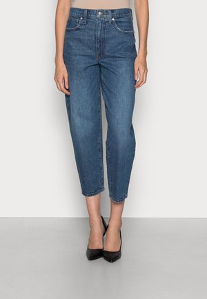 Kobieta nosząca wysokie, skrócone niebieskie dżinsy z denimu z zapięciem na guzik i zamek błyskawiczny, w połączeniu z czarnymi szpilkami z zaostrzonym noskiem.