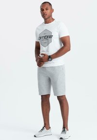 Maglietta bianca con grafica esagonale nera, abbinata a pantaloni corti grigi a righe. Presenta un orologio e scarpe da ginnastica bianche. Design e colori semplici.