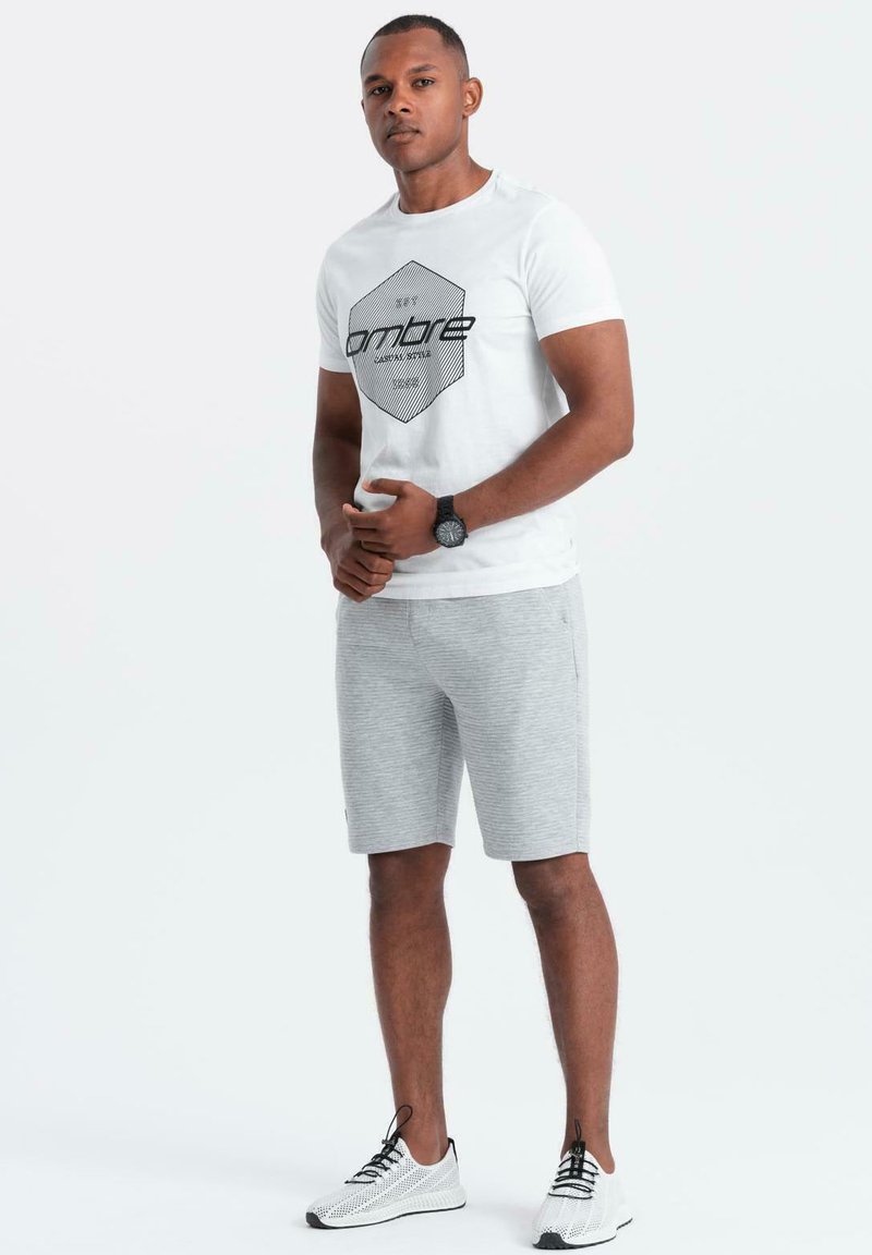 Maglietta bianca con grafica esagonale nera, abbinata a pantaloni corti grigi a righe. Presenta un orologio e scarpe da ginnastica bianche. Design e colori semplici.