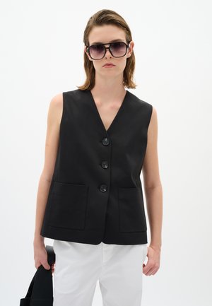Donna che indossa occhiali da sole neri oversize, gilet nero senza maniche con bottoni e tasche, pantaloni bianchi, che tiene una borsa nera su sfondo bianco.
