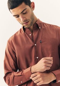 Chemise boutonnée marron en tissu lisse, avec une poche poitrine, un col à boutons et des boutons blancs. Coupe ajustée avec manches longues.