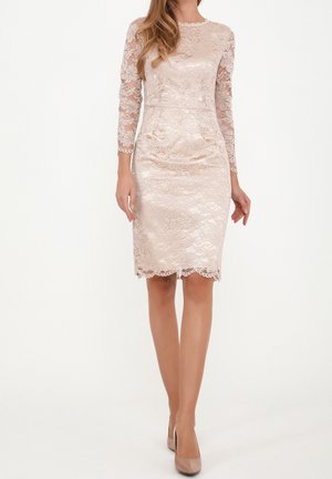 Femme portant une robe en dentelle rose clair, longueur genou, manches longues, avec des talons assortis, debout sur un fond blanc.