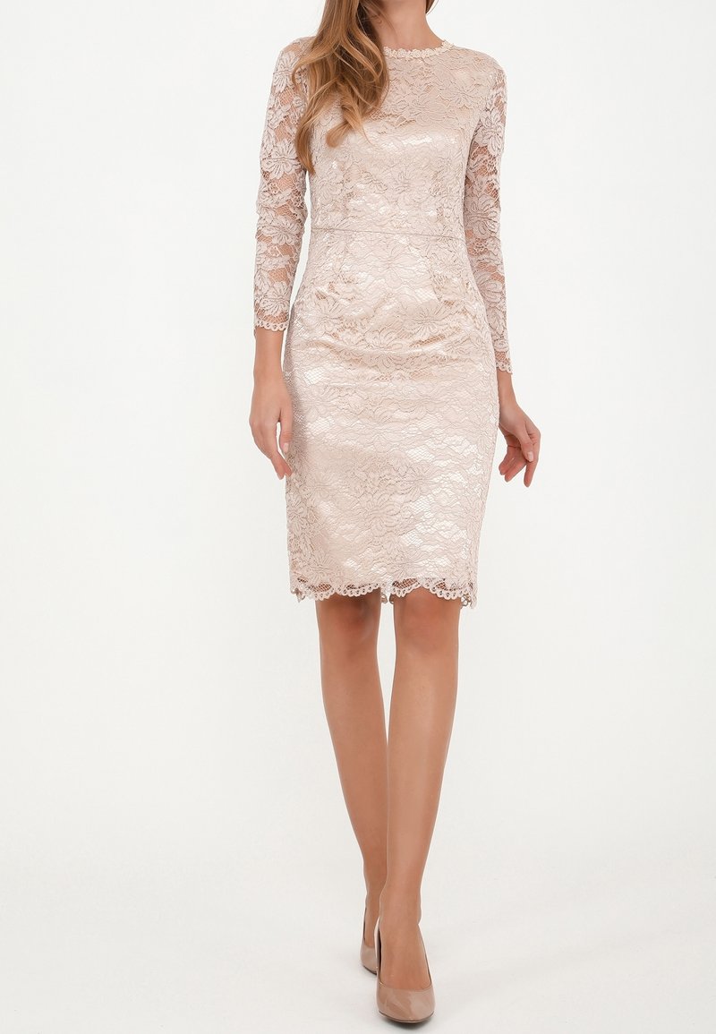 Femme portant une robe en dentelle rose clair, longueur genou, manches longues, avec des talons assortis, debout sur un fond blanc.
