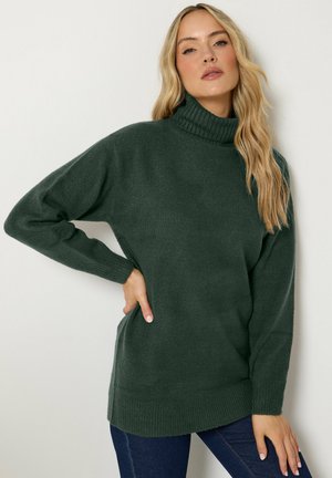 Femme blonde portant un pull à col roulé vert foncé et un jean bleu, debout avec une main sur la hanche sur un fond uni.