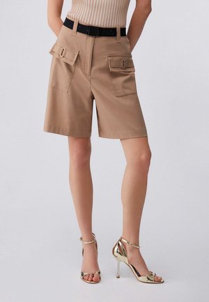Donna che indossa pantaloncini cargo beige al ginocchio con tasche a patta e una cintura nera, abbinati a sandali dorati con tacco alto.