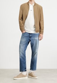 Chaqueta bomber color caqui, polo blanco con logo, jeans azul desgastado y zapatillas beige. El conjunto presenta puños acanalados y un ajuste relajado.