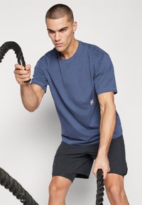 Tricou din bumbac de culoare bleu marin, cu maneci scurte și un material texturat, având un mic logo pe partea stângă, asortat cu pantaloni scurți sport negri.