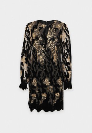 Un modèle se tient devant un fond uni, portant une robe noire aux genoux ornée de broderies florales dorées et des chaussures noires à talons.