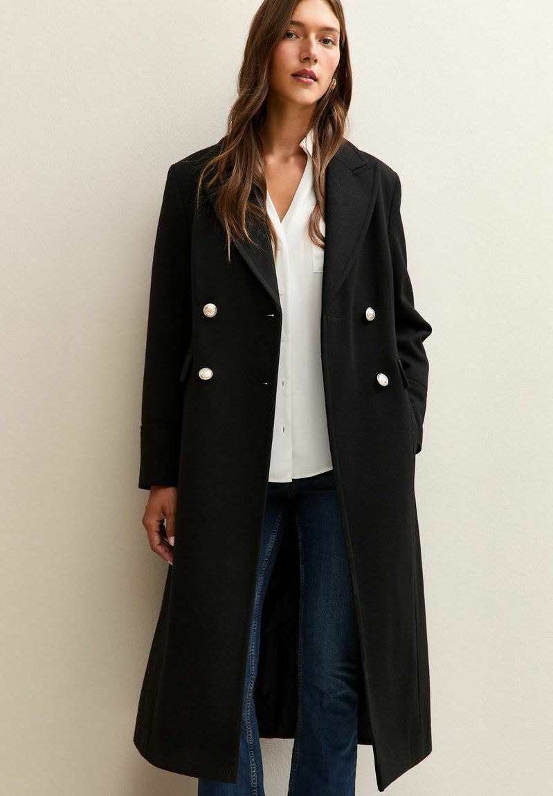 Kvinna iklädd en lång svart dubbelknäppt kappa med pärlknappar, vit blus och blå jeans, stående mot en ljus vägg.