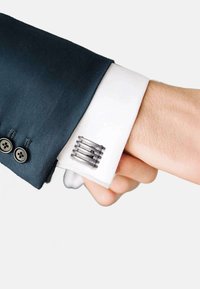 Silver cufflink on a white dress shirt cuff, featuring a modern, geometric design with textured, horizontal lines and a dark contrast against navy fabric.
