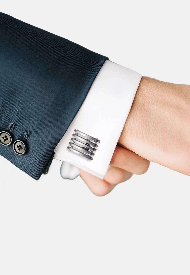 Silver cufflink on a white dress shirt cuff, featuring a modern, geometric design with textured, horizontal lines and a dark contrast against navy fabric.