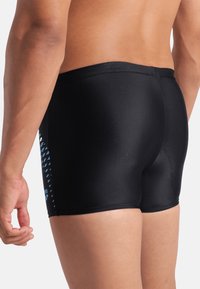Pantaloni da nuoto neri realizzati in tessuto liscio, con un motivo geometrico blu sul lato sinistro. Vestibilità su misura con vita alta.