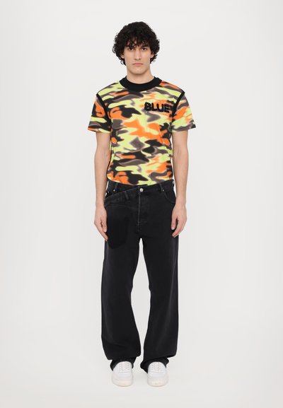 Korte mouwen camo t-shirt in oranje, geel en groen; zwarte accenten; "BLUE" tekst; gecombineerd met zwarte brede jeans en witte sneakers.