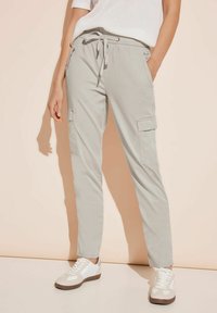 Codzienne jasnoszare spodnie cargo z regulowanym pasem na troczki, noszone z białymi trampkami i białym krótkim topem na krótki rękaw, ręce w kieszeniach.