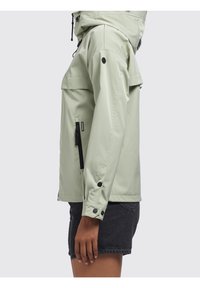 Jasnozielona wodoodporna kurtka z kapturem, kieszenią na zamek, zatrzaskami na mankietach oraz luźnym krojem. Gładka, błyszcząca faktura.