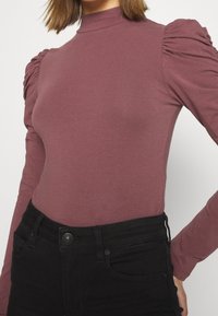 Długi rękaw w kolorze mauve z wysokim dekoltem i marszczonymi puff sleeves, wykonany z miękkiego materiału. Połączony z czarnymi dżinsami o wysokim stanie.