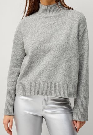 Vrouw draagt een lichtgrijze geribbelde coltrui en glanzende zilveren broek tegen een effen achtergrond.