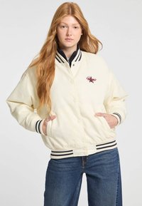 Crème-kleurige bomberjack met gestreepte marineblauwe accenten op de kraag en manchetten, met een logo aan de voorkant en zijzakken. Textuurstof.
