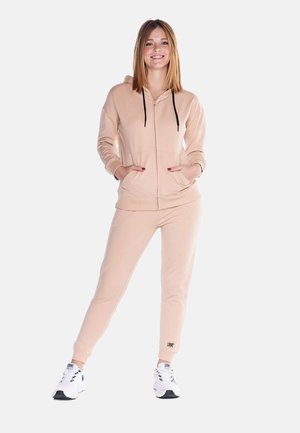 Une femme souriante porte un sweat à capuche rose pâle zippé et un pantalon de jogging assorti, avec des baskets blanches, sur un fond uni.
