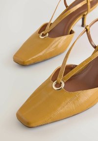 Geltoni tekstūruoti bateliai su atvira kulne, kvadratine nosyte, plonu kulnu ir auksiniu, apvaliu metalo akcentu ant dirželių. Lygios odos apdaila.