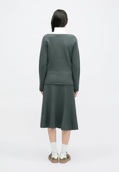 Σκούρο πράσινο πλεκτό πο�υλόβερ και αντίστοιχο μίντι φούστα, με υφή. Συμπληρώνεται με λευκές κάλτσες μέχρι τον αστράγαλο και ελαιόχρωμα αθλητικά παπούτσια.