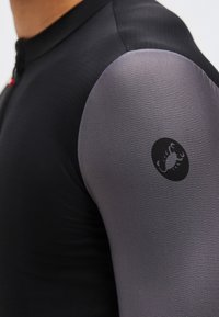 Δεξί χέρι ατόμου που φοράει αθλητική μπλούζα σε μαύρο και γκρι χρώμα με μαύρο λογότυπο σκορπιού στο γκρι μανίκι.