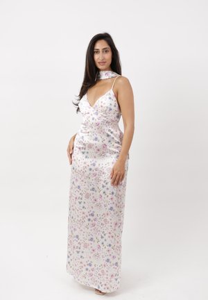 Donna con capelli lunghi e scuri che indossa un abito maxi bianco senza maniche con stampa floreale e scollo a choker, in piedi su uno sfondo bianco uniforme.
