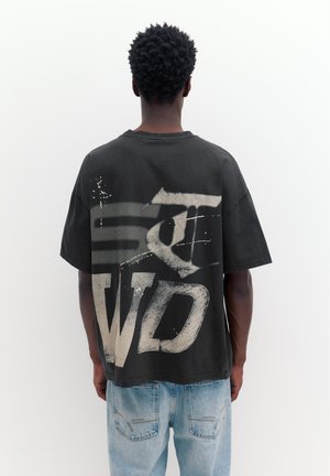 Személy fekete, túlméretezett pólóban, amelyen nagy fehér grafikai nyomat található, és világoskék farmernadrágot visel, hátulnézetben, egyszínű fehér háttér előtt.