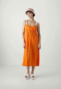 Vestido maxi sin mangas de color naranja con escote fruncido, tirantes blancos y aberturas laterales. Combinado con sandalias de plataforma blancas y un sombrero de cubo a rayas.